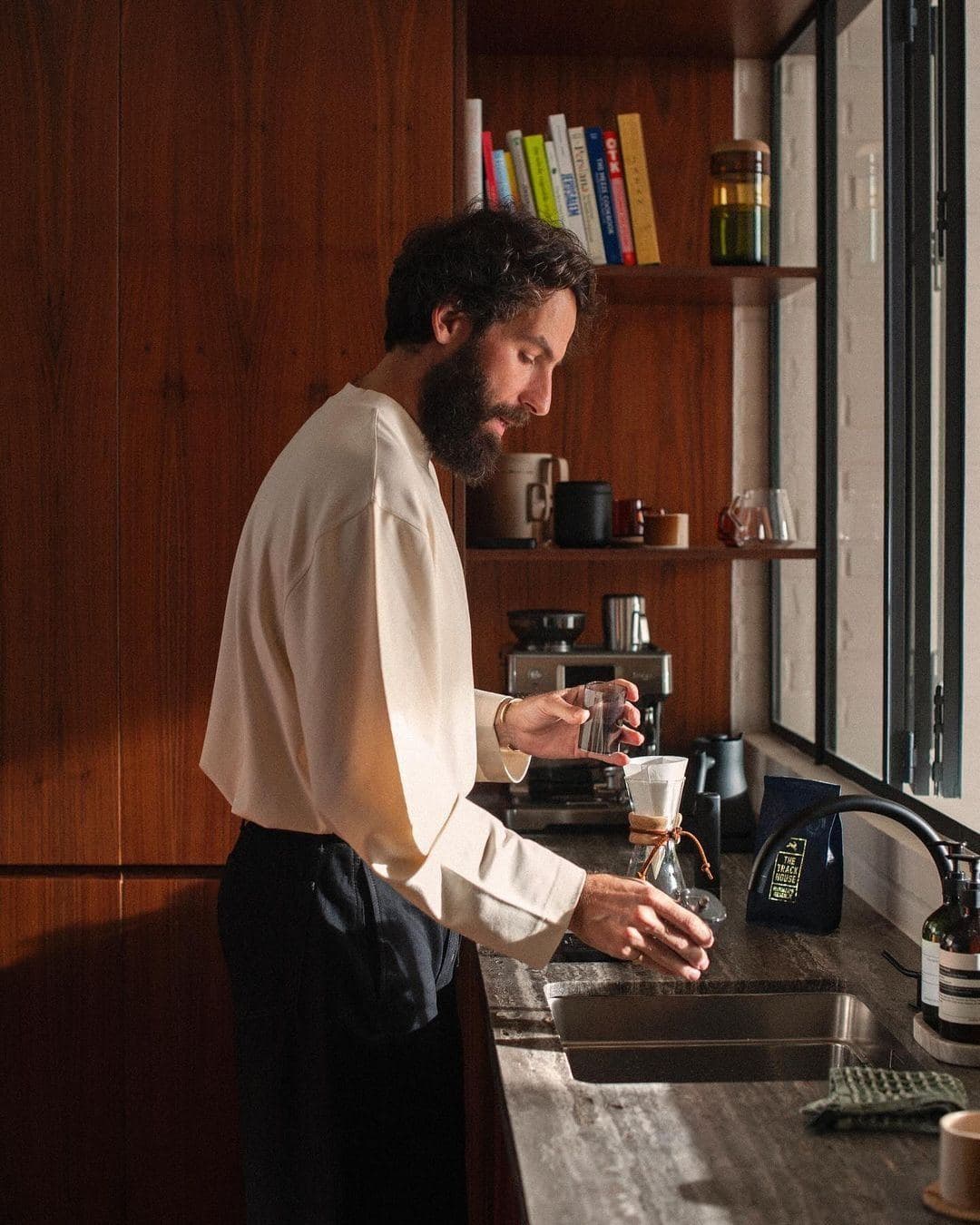 A man making pour-over coffee at a kitchen counter in morning light